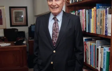 Retrato de Carlos Denton, fundador de CID Gallup, pionero en estudios de opinión pública y consultoría política en América Latina, posando en su oficina junto a una biblioteca.