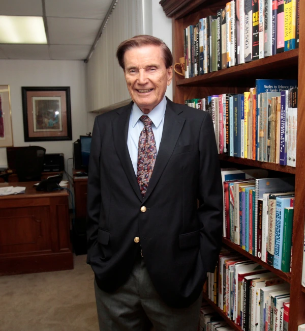 Retrato de Carlos Denton, fundador de CID Gallup, pionero en estudios de opinión pública y consultoría política en América Latina, posando en su oficina junto a una biblioteca.